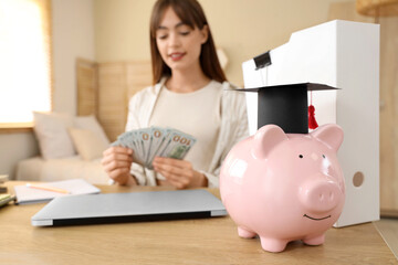 Piggy bank with graduation hat on table against female student at home, closeup. Education savings...