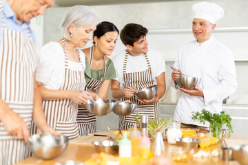 Smiling middle-aged man cook conducting culinary classes for learners standing around kitchen table...