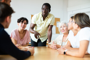 Fototapeta premium Positive friendly African American English tutor giving language courses to group of interested adults of different ages and nationalities sitting around table in classroom..