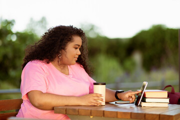 A young woman sits at a wooden table in a serene outdoor setting, sipping coffee from a cup while...