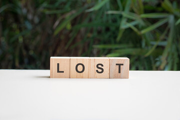 Lost, word written on square wood block, on top of white table with palm plant at the background.