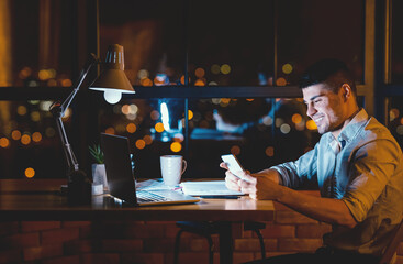 Business Chat. Man Using Mobile Phone Texting Sitting At Laptop In Modern Office In The Evening. Copy Space