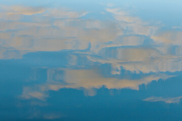 A peaceful reflection of soft clouds floating on the calm surface of a blue lake. The abstract shapes and muted colors evoke a serene, tranquil atmosphere.