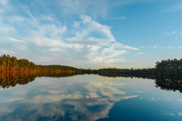 A serene landscape featuring a crystal-clear lake reflecting the blue sky, white clouds, and...
