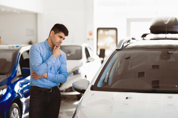 In Doubt About Buying Car. Pensive man looking at auto standing in dealership showroom. Free space for text