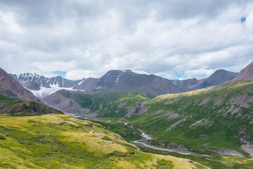 Wide green alpine valley with snake river among hills and rocks with vast view to large snow mountain top and big glacier tongue under cloudy sky. Serpentine river in high mountains. Shadows of clouds