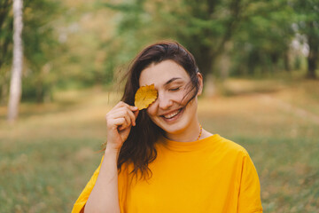 A woman enjoys a sunny autumn day while playfully holding a yellow leaf in a park, surrounded by colorful foliage