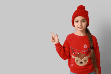 Cute little girl wearing Christmas sweater pointing at something on grey background