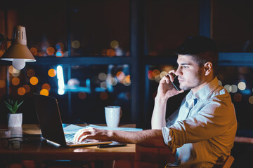 Overnight Work. Businessman At Laptop Talking On Phone Sitting Near Panoramic Window With Night City View In Office. Empty Space