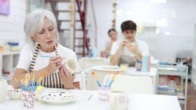 Interested elderly woman attending pottery class, enthusiastically painting on ceramic cup, creating unique custom design. Creative hobby concept