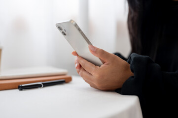 A close-up of a woman in a black sweater using her smartphone while sitting at a table indoors.