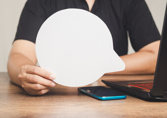 Man holding a blank speech bubble at a desk with laptop and smartphone.