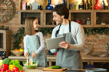 Young Dad Looking Recipe In Digital Tablet While Cooking With Little Daughter At Home, Having Fun Together In Kitchen, Preparing Healthy Lunch, Making Vegetable Salad, Enjoying Homemade Food