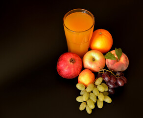 A glass of a mixture of fruit juices on a black background, next to ripe apples, orange, grapes, pomegranate and peach.