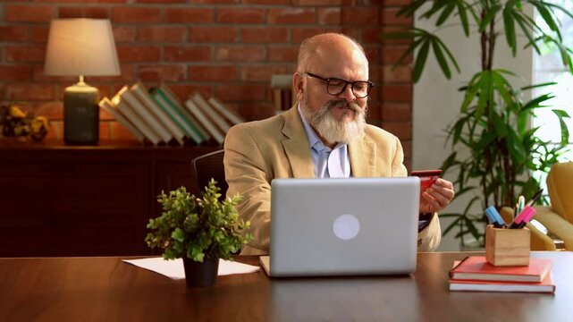 Indian Asian elderly businessman with a beard, dressed in a suit, sitting in a modern office, using a debit or credit card to make online purchases through a laptop