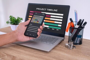 Male hand using a calendar app on smartphone to check the project timeline, milestones and planning progress on laptop