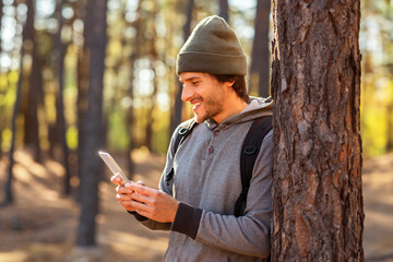Happy young man with backpack chatting with friends on phone in forest, copy space