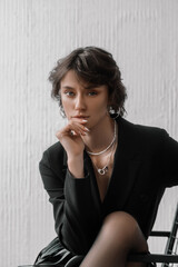 In a stylish indoor setting, a woman with short, wavy hair wears a black blazer and is deep in thought, resting her chin on her hand while maintaining a confident expression