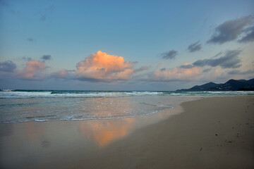 A scenic view of a calm ocean with gentle waves lapping on a sandy beach at sunset. A single large, puffy cloud floats in the sky, reflecting its soft pink hue onto the water