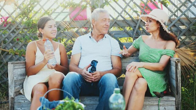 European woman in straw hat sitting on wooden bench, chatting sweetly with sister and elderly father. Rustic setting, young women and gray-haired man on background of wooden trellis discussing news