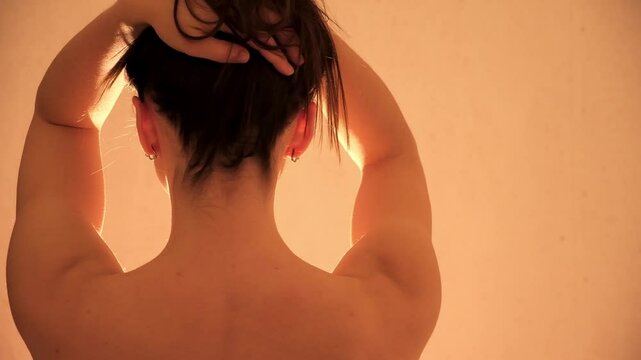 Young 30s sexy woman rotates head warmuping neck. Back view. Morning stretching. The girl holds her long dark hair with hands, showing neck and shoulders. Evening body bliss ritual. Arousal and lust.
