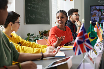 Diverse group of students sitting at table with country flags and chatting during international conference in school classroom