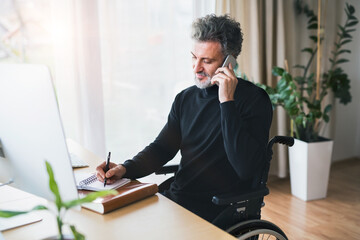 Naklejka premium Handsome man in wheelchair working from home office, making phone call and writing notes in planner. Telecommuting and home office.
