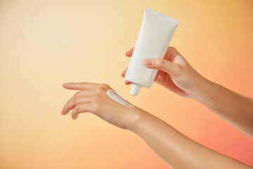 The female model gently squeezes a sample of cream onto her other hand from an unlabeled white tube under a bright orange background, suitable for a sunscreen advertisement.