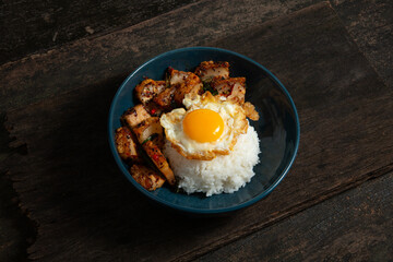 Stir-fried crispy pork with fried egg on rice in blue bowl on old wooden table
