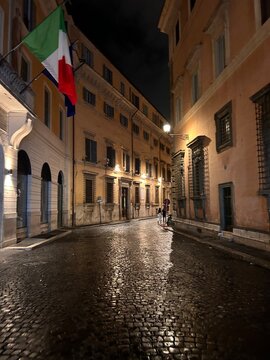 A  charming night scene of a quiet street in Rome, Italy, illuminated by warm street lights reflecting on the wet cobblestone pavement. The Italian flag is visible on a nearby building, adding a patri