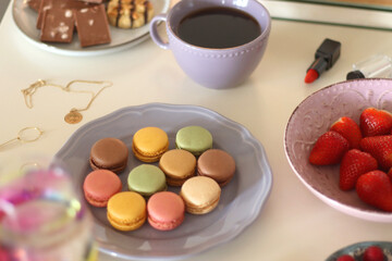 Plate of pastel macarons, cookies and chocolate, cup of tea of coffee, glass of bubble water, various berries, books and accessories on the table. Selective focus, pastel colors.