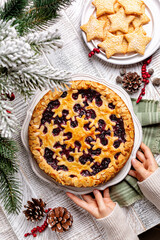 Winter Christmas or New Year festive bakery. Hands holding baked mixed berry almond crust pie or tart with snawflakes decoration. Homemade star shaped cookies on a white table, vertical image.
