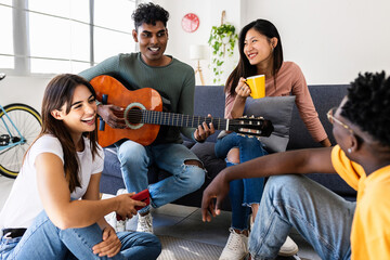 Young group of millennial people having fun together at modern flat. Happy indian man playing the guitar enjoying time with his friends at home