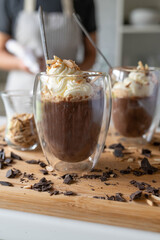 Homemade chocolate pudding with whipped cream topping in glasses served by a woman with apron in the kitchen