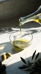An olive oil being poured into an empty glass bowl, surrounded by olives and kitchen utensils on the side, set against a light gray background