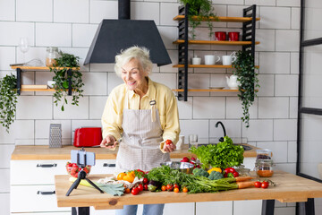 happy beautiful elderly gray haired senior woman cook in cozy kitchen with fresh organic vegetables, records a recipe on phone camera as food blogger or cooks according to recipe online and smiling