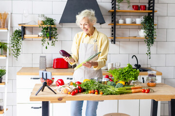 happy beautiful elderly gray haired senior woman cook in cozy kitchen with fresh organic vegetables, records a recipe on phone camera as food blogger or cooks according to recipe online and smiling