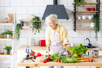 happy beautiful elderly gray haired senior woman cook in cozy kitchen with fresh organic vegetables, records a recipe on phone camera as food blogger or cooks according to recipe online and smiling