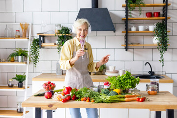 happy beautiful elderly gray haired senior woman in headphones cook in cozy kitchen with fresh...