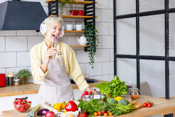 happy beautiful elderly gray haired senior woman in headphones cook in cozy kitchen with fresh organic vegetables, having fun and singing
