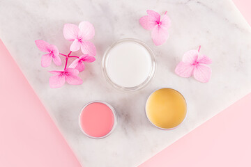 Three cosmetic jars with natural lip balm in different colors on a marble stand with pink flowers. top view. Natural care. Care and beauty concept.