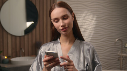 Caucasian beautiful woman female lady girl in home hotel bathroom pensive scrolling mobile phone app shopping online choose cosmetics order delivery using smartphone purchase gadget addict internet