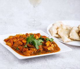 A hearty dish of tender chicken curry with bright spices, garnished with fresh cilantro, served alongside soft, warm naan bread on a white plate. served with a glass of chilled white wine.