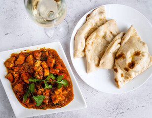 A hearty dish of tender chicken curry with bright spices, garnished with fresh cilantro, served alongside soft, warm naan bread on a white plate, served with a glass of chilled white wine.