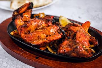 A vibrant, sizzling platter of tandoori chicken served on a wooden board, garnished with lemon slices in the background.