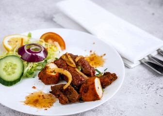 A well-plated dish featuring grilled meat chunks, garnished with spices and served alongside a fresh salad. The salad includes cucumber slices, a red onion ring, and vibrant tomatoes.