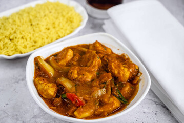 Delicious plate of chicken curry was served in a white bowl, showcasing tender chicken pieces in a rich, spicy sauce with visible vegetables and herbs. A side of fluffy yellow rice enhances the dish