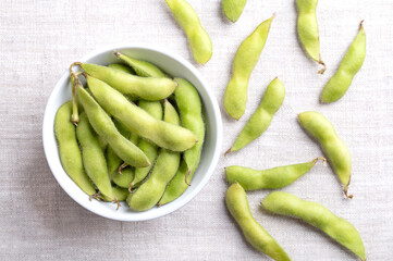 Fresh edamame, green soybeans in the pod, in a white bowl, on linen. Raw, immature soybeans, Glycine max, used boiled or steamed and salted, as a common side dish and appetizer in Japanese cuisine.