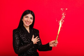 A brunette in a shiny dress in a good mood poses with a sparkler in the studio. A model on a red background looks into the camera