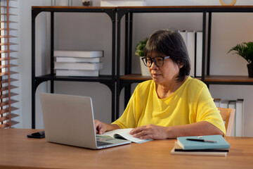 Senior asian woman reading a book and study while laptop on desk office in living room at home, elderly woman retirement learning education with concentration, lifestyle and education.